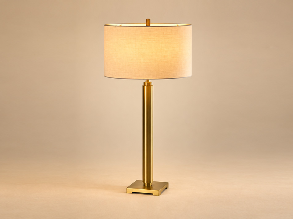Tom brushed gold metal table lamp with tall cylindrical column, square base and white linen drum shade, designed for contemporary living rooms and console tables.