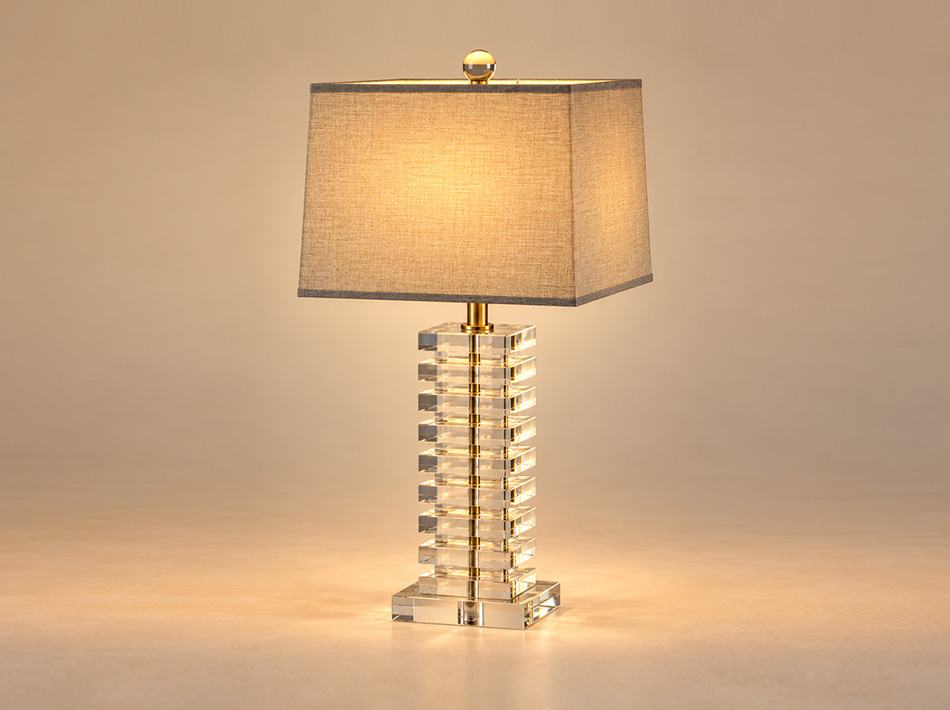 Halston brushed gold table lamp featuring a tall column of stacked clear crystal elements, geometric detailing and a square light brown linen shade.