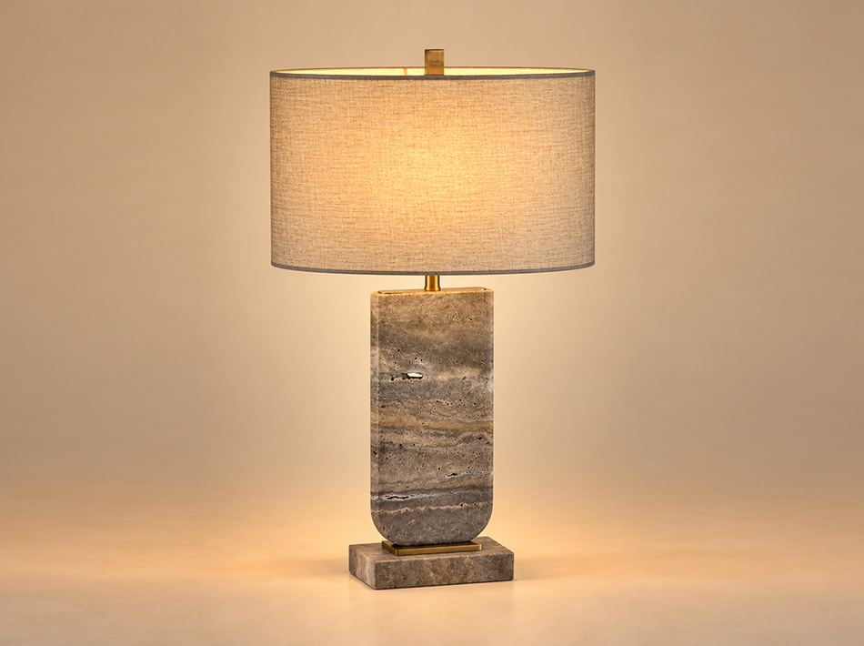 Maple brushed gold table lamp with a tall grey Silver Travertine stone base, natural horizontal veining and oval light brown linen shade.
